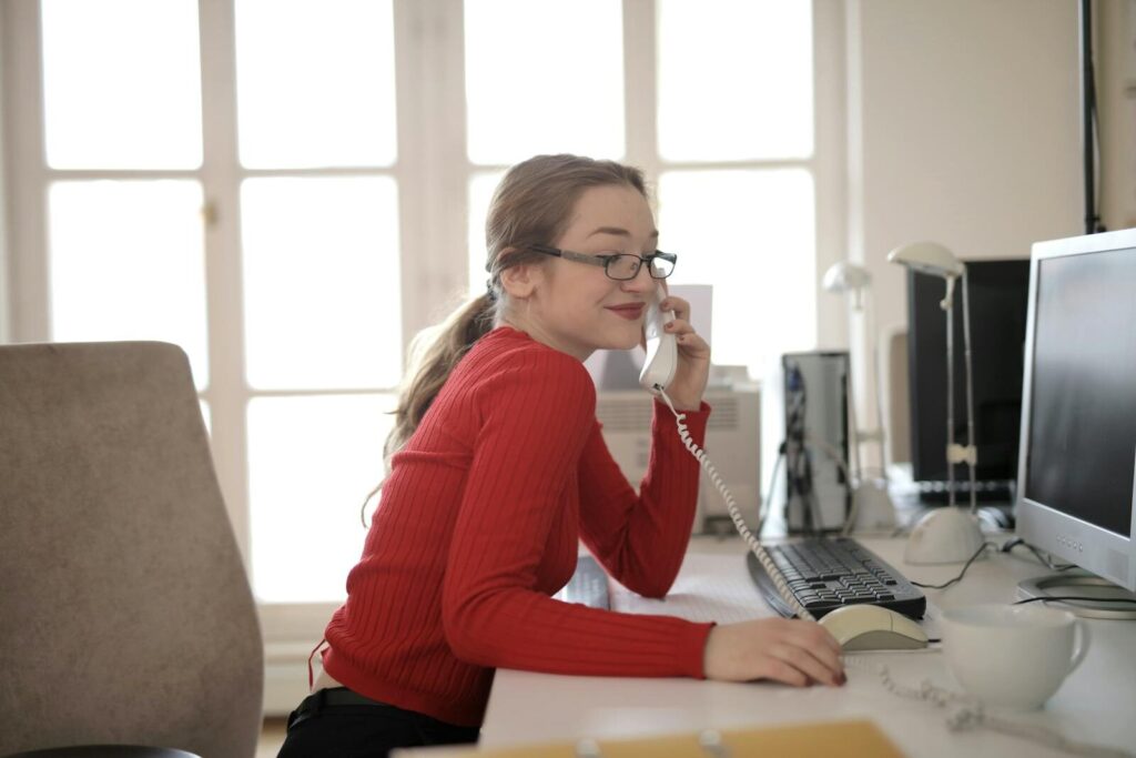 Reduced Expenses and Fair Billing 1 Caucasian woman in red sweater talking on landline phone at workplace in natural light.