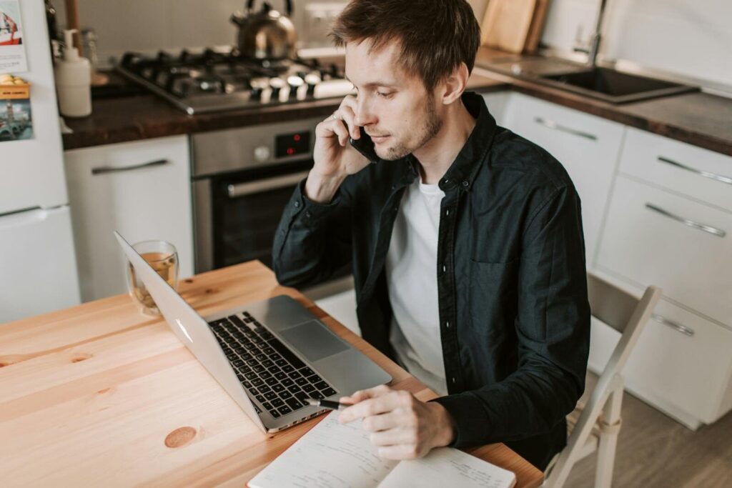 Young male remote worker in casual wear talking on smartphone and watching netbook while sitting with pen and open notebook at wooden table in apartment